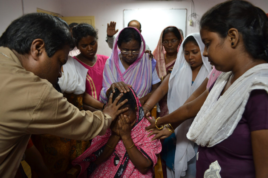 Indiska kvinnor och män lägger händerna på en kvinna i mitten i ett mindre rum.